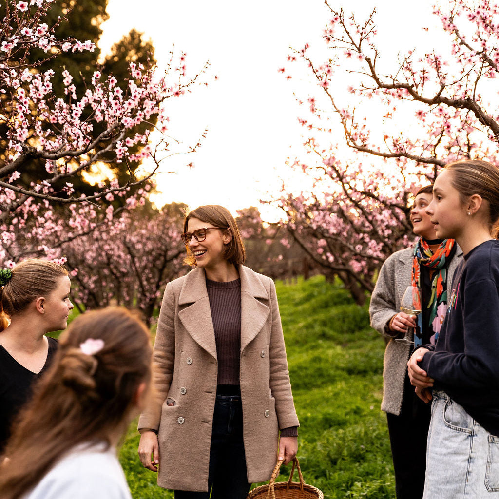 A woman in a beige double-breasted coat holding a wicker basket stands in a grassy orchard with blooming pink trees, surrounded by 4 people on the blossom tour.